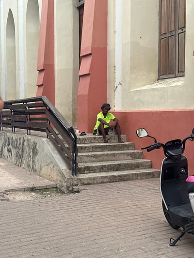 In the Perímetro Urbano Barranquilla, Colombia, a person wearing a bright yellow t-shirt and dark shorts sits on the concrete steps of a building. The person has on a dark cap and appears to be resting, with their hands clasped and feet bare. The steps are part of a staircase that leads up to the building's entrance, which features a reddish-orange facade and tall, cream-colored architectural elements with arched openings. A dark wooden railing runs alongside the stairs. In the foreground, a black and white scooter is parked on a brick-paved sidewalk. The environment appears urban, with some signs of wear on the building's exterior and the surrounding pavement.