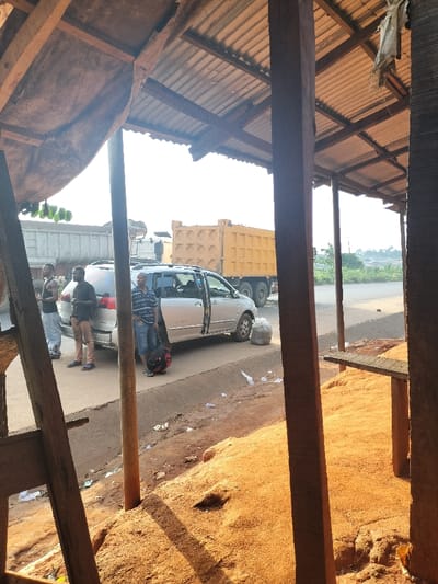 This image depicts a roadside scene in Oluku, Nigeria. The foreground shows a partial view of an open-sided wooden structure with a corrugated metal roof, supported by timber posts and rafters. A raised bank of reddish-brown soil, consistent with laterite, forms the immediate ground.
In the midground, a paved road with dirt shoulders runs left-to-right. On the left side of the road, a silver minivan, possibly a Toyota Sienna, is stopped with its passenger-side sliding door open. Three adult males are standing near the minivan. From left to right: one male in a dark t-shirt and light pants, another male in a dark t-shirt and light brown pants, and a third male in a striped polo shirt and blue shorts, holding a black backpack. A grey plastic bag rests on the ground near the front passenger tire of the minivan. Scattered white litter is visible on the dirt shoulder of the road.
Behind the minivan, a white dump truck with a full, grey load in its bed is partially visible. Further down the road to the right, a large yellow dump truck is parked or moving slowly. The background features a hazy, vegetated landscape under a bright, clear sky, indicating daylight conditions with direct sunlight.