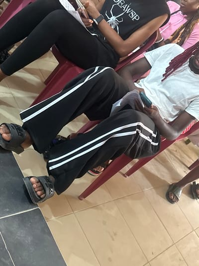 The scene, located in Iriebe, Nigeria, is an indoor, eye-level shot capturing portions of at least five individuals seated on maroon plastic chairs. The floor is tiled, with dark grey tiles on the left and light brown/beige tiles on the right, separated by a linear grout line.

In the left foreground, an individual's legs are visible, clad in black athletic pants with two parallel white stripes running down the outer seams. Their feet are covered by dark grey slide sandals, exposing toes.

Behind this individual, another person is partially visible, wearing black leggings and a black t-shirt featuring a white spider graphic and text "BAWSP." A black smartwatch is present on their left wrist. Their hands hold a light-colored smartphone and white papers.

To the right, a person with reddish-brown braided hair wears a white t-shirt and black pants, also featuring two parallel white stripes. This individual holds a dark-colored smartphone and papers on their lap.

Further to the right, a pink garment and an arm indicate another seated person, largely obscured. In the lower right corner, the feet of a fifth individual are visible, wearing dark slide sandals.

Individuals are generally passive, seated, with at least two observed interacting with their mobile devices and papers. The lighting suggests an indoor setting.