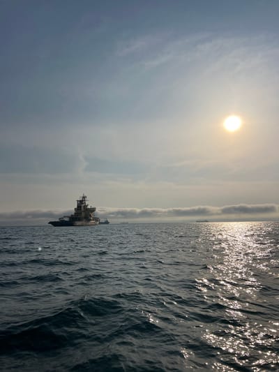 The image depicts a ship at sea under a bright sun. The sea is choppy, reflecting sunlight. The sky is hazy with clouds. The ship is the focal point, appearing to be a large vessel, possibly a military or cargo ship. The image was not taken in Novomoskovsk, Russia as this is a landlocked city.