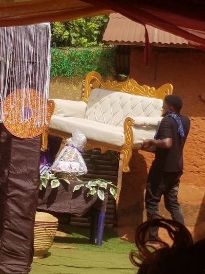 The scene is set outdoors in Ochadam, Nigeria. A male subject, dressed in a dark t-shirt and dark trousers, with a blue patterned fabric draped over his left shoulder, supports a large, ornate sofa featuring a gold-colored frame and cream-colored tufted upholstery. To the left of the sofa, a decorative arrangement includes a white vertical string curtain from which a round, orange, gold, and purple mosaic-patterned disc hangs. Below this, a table covered with a dark brown fabric is adorned with green artificial ivy. On the table rests a basket containing an item wrapped in clear cellophane and tied with a purple ribbon. A light brown woven basket is on the green artificial turf in the lower-left foreground, adjacent to a dark blue cylindrical object. The background consists of an earth-toned wall topped with green foliage and a section of a corrugated metal roof. Overhead, sections of brown and red fabric drapes indicate a temporary canopy. The upper portion of a head with dark, curly hair is partially visible in the lower-right foreground.