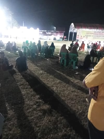 The image depicts an outdoor evening event in Alueanou Barat, Indonesia, likely a community gathering or religious ceremony. Numerous individuals are seated on the ground and on chairs in a field, with some wearing headscarves. In the background, a tent with a banner is visible, suggesting an organized activity. The lighting appears to be artificial, casting shadows across the grassy terrain. A structure resembling a building with lights is also present in the distance. The overall scene suggests a low-key, communal event taking place after dark.