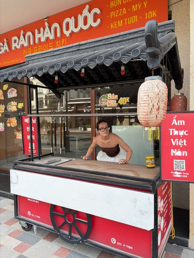 A young woman with glasses and dark hair, wearing a brown tube top and white skirt, stands behind a food cart under an overhang with a Korean-inspired roof. She is leaning forward, with her hands on the cart's counter. The food cart is red and white, with black accents and wheels. On the cart, there are various signs with Vietnamese and Korean text. One sign on the cart's side reads "K22. K-TOWN". The overhead sign displays "GA RÁN HÀN QUỐC" and Korean text. The background shows a street with buildings and trees. The scene is outdoors, likely in a commercial area.