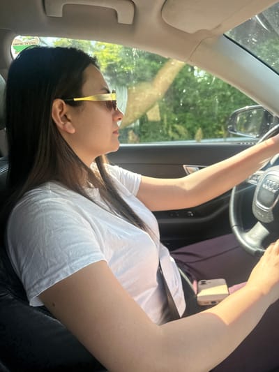 Inside a vehicle, located in Ohoden, Bulgaria, a female driver is depicted from the right side. She has long, dark hair, wears a white short-sleeved t-shirt, dark pants, and gold-framed Bvlgari sunglasses. A dark mole is visible on her upper left arm. She is secured by a seatbelt across her chest and lap. Her hands are positioned on a black steering wheel, which bears the Audi logo. A light-colored smartphone is visible, placed beneath her left thigh/lap. Through the windshield and driver's side window, bright daylight illuminates green foliage. The windshield exhibits faint smudges and a reflected, inverted image of text in its upper left quadrant.