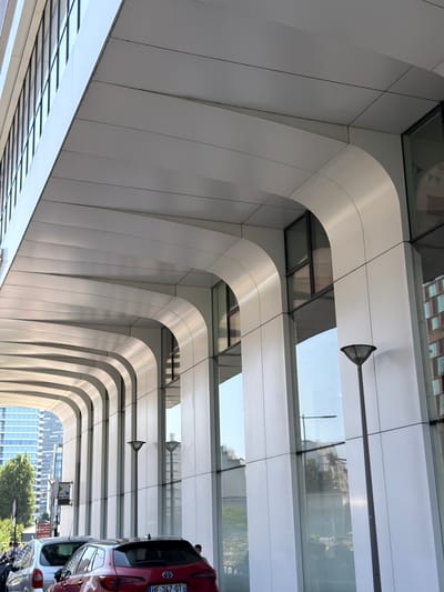 The image depicts a modern building in Nanterre, France, characterized by its sleek, white arched overhang and large glass windows. The architecture suggests a commercial or office building with a covered walkway or arcade along its facade. The overhang is composed of curved, white panels with visible seams, extending horizontally and creating a repeating pattern of arches. Below the overhang, the building features extensive glass paneling, reflecting the surrounding environment and sky.
Several streetlights with distinctive dark, mushroom-shaped lamps are visible along the walkway. In the foreground, cars are parked, indicating a public area or street. A red hatchback and a silver hatchback are clearly visible, with parts of other vehicles and a person on a bicycle further back. The background shows other buildings, including a taller, modern skyscraper and older buildings with brick facades, along with trees and greenery, suggesting an urban setting with varied architectural styles. The sky is bright, implying daylight. The overall scene is one of a contemporary urban environment in Nanterre.