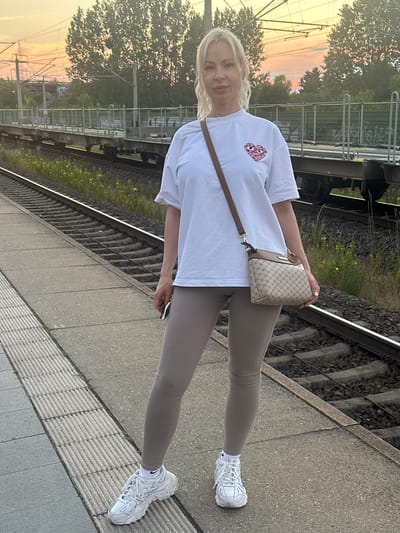 A blonde woman stands on a train platform in Wustermark, Germany, near a railway track during sunset. She is wearing a white t-shirt with a pink heart graphic, beige leggings, and white sneakers. A brown crossbody bag hangs from her shoulder. She is holding a dark object, possibly a phone, in her left hand. The background shows train tracks, a train, trees, and a sunset sky with power lines.