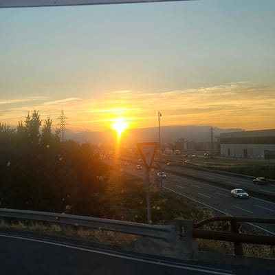 The image captures a sunset scene on a highway near Gurb, Spain. The sun, a brilliant yellow orb, is low on the horizon, casting a warm, golden light across the sky and the surrounding landscape. Wispy clouds are scattered across the sky, tinged with orange and yellow hues.
In the foreground, a concrete barrier with a rusty metal railing runs along the edge of the road. A dark asphalt road stretches into the distance, with multiple lanes of traffic. Several cars, illuminated by headlights and the sunset, are driving on the highway.
To the left of the highway, a dense cluster of dark green trees forms a thick foliage. Behind the trees, a tall, lattice-like electrical pylon stands prominently against the sky. In the background, mountains are visible in the hazy distance, silhouetted against the bright sunset. On the right side of the highway, industrial buildings with large, flat roofs are situated. A triangular road sign, depicting a circular symbol with a line through it and a smaller circular symbol below, is visible on the side of the road. The overall scene conveys a sense of transition, with the natural beauty of the sunset juxtaposed with the man-made elements of the highway and industrial area.