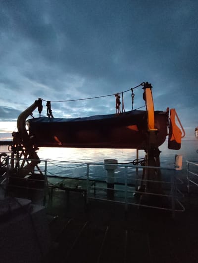 The image depicts a lifeboat mounted on davits aboard a vessel at dusk. The sky is overcast with hues of blue and gray, and the water appears calm with a subtle reflection of the fading sunlight. The lifeboat is dark and appears to be covered, with an orange safety device visible at its stern. The davit system, made of metal, is positioned above the lifeboat, with cables and pulleys used for deployment. The deck of the vessel is visible in the foreground, showing a dark, textured surface and railings. The scene suggests a maritime setting, potentially in the vicinity of Shcheleiki, Russia, given the context, but the exact location is not visually confirmed within the image itself. No people are present in the frame.