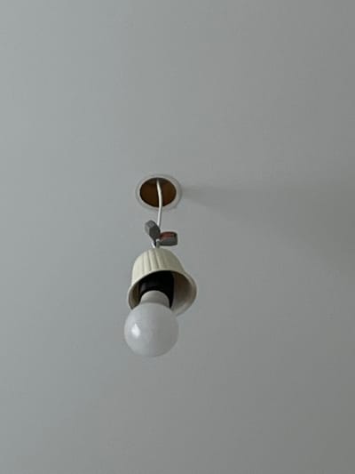 An indoor scene in Zainsk, Russia, depicts a partially dislodged or damaged ceiling light fixture against a plain, light-colored ceiling. A spherical white light bulb is screwed into a visible black socket. Above the socket, a cream-colored, bell-shaped light shade with vertical ridges hangs unevenly, exposing the internal components. White electrical wiring descends from a circular opening in the ceiling, revealing the brown edge of the aperture. Two small, grey electrical wire connectors are visible on the wiring segment above the shade, one featuring a red marking. No individuals are present in the frame.