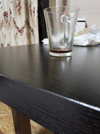 This is a medium-angle, close-up shot of a dark wooden table in what appears to be a home setting. A clear glass mug with a handle is positioned on the table, containing a small amount of dark liquid at the bottom, likely coffee or tea. The mug is partially in focus. In the background, a light-colored curtain with a floral pattern featuring burgundy and beige tones is visible. To the right of the mug, a white plastic package, possibly of wipes or a similar product, is present, along with some crumpled paper or fabric. The overall lighting suggests an indoor environment, possibly during the daytime, but there are no strong indicators of the specific time of day or weather conditions. The scene is static, with no activity taking place. The focus is on the still life arrangement of the mug and the table. There is no visible text in the image. The context of Brish, Russia, does not provide specific visual cues within the image itself.