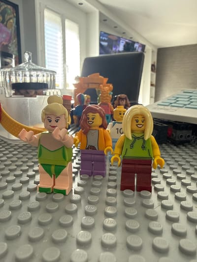 This image depicts a static arrangement of LEGO minifigures set up on a light grey studded baseplate within a residential interior in Limeil-Brévannes, France.
In the foreground, three female minifigures are positioned upright. From left to right:
1. A minifigure with blonde hair in a bun, light pink skin, wearing a two-piece green swimsuit. Her arms are slightly raised, and she has a smiling facial expression.
2. A minifigure with auburn, wavy hair, light pink skin, wearing an open orange jacket over a grey shirt with a pendant, and dark purple pants. She also displays a smiling expression.
3. A minifigure with long blonde hair, light pink skin, dressed in a lime green hooded sweatshirt and dark red pants. This figure also has a smiling expression.
In the mid-ground, several other LEGO minifigures are present in softer focus. These include a male minifigure wearing a white shirt with the number "10" printed on it, and various other figures with different hair colors and outfits. An orange arched LEGO structure is visible behind the central group of minifigures.
The background reveals a brightly lit indoor environment. On the left, a large window with white horizontal blinds is visible, allowing natural light into the scene. A clear, multi-tiered glass object, possibly a decorative container, stands behind the leftmost minifigure. In the center-right background, a large, dark, rectangular object, consistent with a television screen or computer monitor, is present. To the far right, a light-colored object with a grid pattern, possibly a keyboard or part of a larger LEGO build, is partially visible. The overall setting is a domestic room.