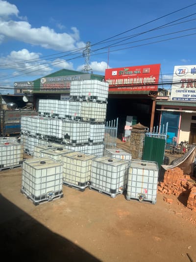 The image shows a stack of intermediate bulk containers (IBCs) in front of two businesses. One sign reads "SY STEEL VINA KOREA COMPANY" and another is "NHÀ MÁY CÁN TÔN-SA." The containers are caged in metal frames. Overhead power lines are visible against a blue sky with clouds. Bricks and other materials are visible beyond the IBCs. The environment appears to be an industrial or commercial area.