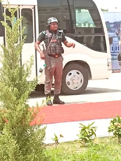 A security officer stands prominently in the foreground, dressed in a dark uniform consisting of a short-sleeved top, tactical vest, pants, and black boots. They wear a dark helmet and have straps across their chest and shoulders, with what appears to be a small white and green emblem or flag hanging from a holster on their right hip. The officer is looking towards the right side of the frame with a focused expression, their right arm slightly raised.

Behind the officer and to their right is a white mini-bus or coaster bus, partially visible. The front section of the bus, including a headlight, part of the windshield, and a wheel, can be seen. Faint reflections on the bus window show what might be inverted text, possibly "HA".

The ground in front of the officer is covered by a red surface, resembling a carpet or painted pathway, which transitions to a lighter, possibly paved, area. In the foreground and to the left, a tall, dense green shrub or tree partially obstructs the view. Smaller green plants and grass are visible at the very bottom of the frame.

The scene takes place outdoors during the daytime, under bright lighting, suggesting clear weather. The officer appears to be on duty, possibly guarding the area or observing. Given the context of Unguwan Sanusi, Nigeria, the uniform and the green/white emblem are consistent with Nigerian security forces.