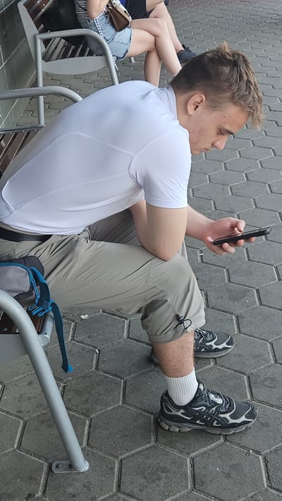 A young man with short, light brown hair and acne on his face sits on a metal bench with wooden slats. He is wearing a white short-sleeved shirt, tan cargo shorts, white socks, and dark athletic shoes with gray and white accents. He is holding a black smartphone in his right hand and looking down at it. A blue strap from a bag is visible over his left thigh. The ground is paved with hexagonal gray stones. In the background, a woman is sitting on the same bench, wearing a denim skirt and a striped top, with her legs crossed. The setting appears to be an outdoor waiting area, potentially a bus stop or train station, in the city of Kysak, Slovakia.