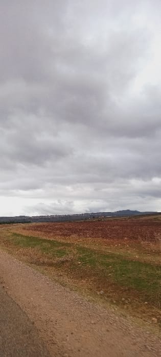 The image displays an expansive, rural landscape under an overcast sky with heavy grey clouds. In the foreground, a light brown dirt path or unpaved road traverses diagonally from the lower left towards the mid-right. Adjacent to the path, the midground consists of agricultural land. To the right, a large section of reddish-brown tilled earth indicates a cultivated field, bordered further right by a strip of green vegetation. To the left of the path, smaller patches of green growth are visible. The distant background features a horizontal line of buildings, likely constituting the city of Ait Naamane, Morocco, positioned on a low ridge. Further to the right in the background, low, dark hills or mountains are visible. No people, animals, specific objects beyond the road and distant structures, or actions are discernible within the scene. The overall environment suggests an agricultural area on a cloudy day.