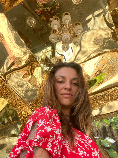 A woman is lying down, possibly on a mirrored surface. She has long brown hair and is wearing a red dress with a white floral print. The surface she is on appears to be made of golden, ornately decorated tiles or panels, reflecting a room with a chandelier, plants, and other furnishings. The location is likely a room within the "Golden Temple" or "Chùa Vàng" at Biconsi Construction Investment Corporation in Dĩ An, Vietnam.