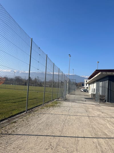 An outdoor scene in Meyrin, Switzerland, depicts a sports facility under a clear blue sky. The image is composed with a chain-link fence running horizontally across the mid-ground, separating a paved area on the right from a lush green field on the left.

In the foreground, the fence stretches from left to right, supported by metal posts, with a small section of an orange structure (possibly part of a goal or net) visible at the bottom left. Beyond the fence to the left, a well-maintained grassy field, likely a football (soccer) pitch, extends into the distance, with a white goal frame discernible further back. Several tall, slender light poles equipped with floodlights are positioned along the fence line, ready to illuminate the field.

Above the field and fence, a vast, unblemished blue sky dominates the upper portion of the frame, with a faint, thin contrail visible high up. In the far distance, beyond the field and trees, majestic snow-capped mountains rise under the clear sky, characteristic of the Swiss landscape.

To the right of the fence, a wide, light-colored concrete or gravel path runs parallel to the field. Further to the right, behind this paved area, industrial or commercial buildings with dark corrugated metal siding are visible, along with some parked vehicles, including a white van or truck.

The scene is bathed in bright daylight, indicating a sunny day, likely morning or afternoon, with no people present, suggesting the facility is currently empty or between activities. There is no visible text in the image.