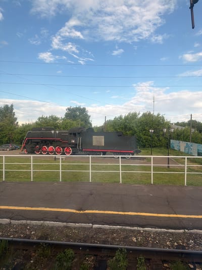 The image shows a stationary black steam locomotive with red wheels displayed outdoors, likely in the city of Vyatskiye Polyany, Russia. The locomotive is positioned on a track, surrounded by a grassy area fenced with white railings. Behind the locomotive, there are trees and some buildings. The sky above is blue with scattered white clouds. Power lines are visible stretching across the sky. The foreground shows a section of a train platform and railway tracks. No people are visible in the image.
