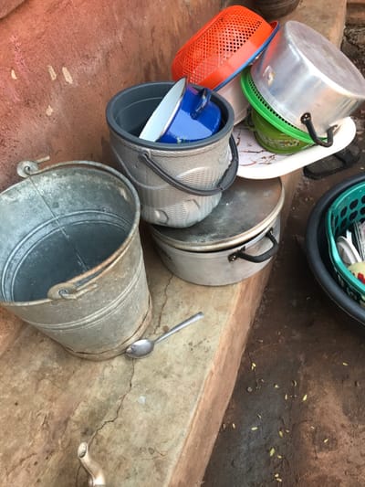 A variety of containers, including pots, baskets, and a metal bucket, are piled on a concrete surface. The pots, made of silver-colored metal, are stacked with lids askew. Bright orange and green baskets are visible. A blue pot sits nested within a larger gray bucket with a black handle. A metal spoon rests precariously on the rim of the metal bucket, which leans against a concrete wall. A tap fixture is on the right side of the image.