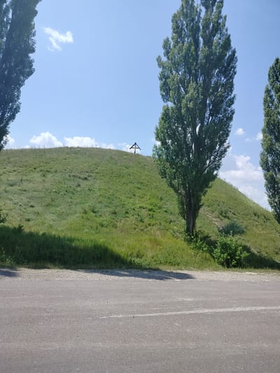 An exterior, eye-level, wide shot captures a scene on a sunny day in Balatina, Moldova. In the foreground, a grey asphalt road with a white stripe runs horizontally across the bottom of the frame. Behind the road, a green grassy slope rises, dotted with sparse vegetation. A tall, thin tree with dark green foliage stands prominently on the right side of the slope, its trunk casting a shadow on the grass. To the left of this tree, another, larger tree with dark green foliage is partially visible. Near the crest of the hill, a small, wooden cross is visible. The sky above is a clear blue, with a few scattered white clouds.