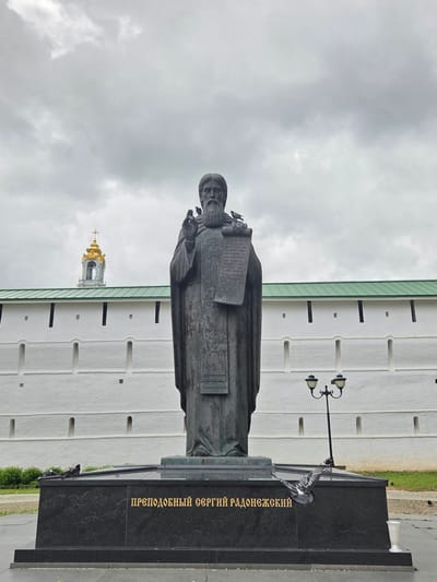 This is a vertically oriented photograph taken outdoors. The primary subject is a large bronze statue of a robed man holding a book or scroll. The statue is situated in front of a white, modern-looking building with a green roof. The building has a series of evenly spaced, arched niches and smaller rectangular windows. To the left of the statue and partially obscured by the building, a golden domed structure, likely a church tower, is visible.

The statue stands on a dark granite pedestal, which has golden text inscribed on its side. The visible text reads "ПРЕПОДОБНЫЙ СЕРГИЙ РАДОНЕЖСКИЙ" (Prepodobny Sergey Radonezhskiy), indicating the statue depicts Saint Sergius of Radonezh. A black lamppost with two lights stands behind the statue.

Several pigeons are present in the scene, with one perched on the pedestal and another on the statue's base. The sky is overcast with diffused light, suggesting a cloudy or possibly rainy day. The overall atmosphere is somewhat somber due to the weather and the dark tones of the statue and pedestal. The image appears to be taken in the daytime. The location context provided is Sergiyev Posad, Russia.
