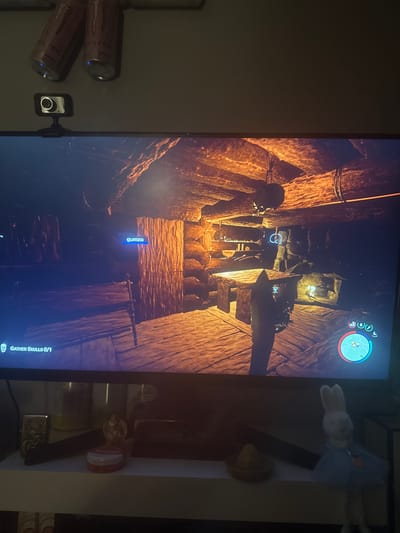 The image depicts an indoor scene captured in Novi Pazar, Bulgaria. A large monitor displaying a video game occupies the central portion of the frame. Above the monitor, two inverted, brown Monster Energy drink cans are affixed to a light-colored wall or ceiling. A black Canyon brand webcam with a silver lens is mounted atop the monitor, slightly to the left.
The monitor displays a dark, rustic video game environment, characterized by rough-hewn wooden logs and planks forming walls and a floor. Warm, orange-toned light sources illuminate parts of the scene, casting long shadows. A skull hangs by a rope from the ceiling beam on the right side of the screen. A wooden workbench or table is positioned in the foreground. In the background, shelves built into the wall hold various items, and a glowing light source is visible. A blue name tag reading "gumza" is present over a wooden structure on the left side of the screen. Game interface elements include "GATHER SKULLS 0/1" in the bottom-left corner and a circular indicator with blue, red, and white segments in the bottom-right corner.
Below the monitor, a white shelf holds several objects. From left to right: a metallic Zippo-style lighter with an embossed design, a small figurine, a circular red and white container, a small bowl containing a yellow lemon-shaped object, and a white plush bunny toy dressed in a light blue tutu with an orange carrot applique. Behind these items, a Glade air freshener can and another generic can are partially visible.