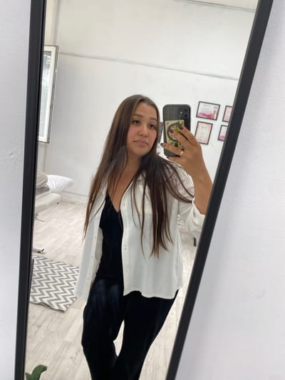 A woman is taking a mirror selfie in what appears to be a room with white walls. She has long brown hair and is wearing a black top with a white open shirt over it. She is also wearing black pants and a gold ring on her right index finger. In the reflection, a light-colored floor with a wooden pattern is visible, along with a grey and white zigzag rug, a white treatment bed with pillows, and framed certificates on the wall. The room seems to be a professional or treatment space, possibly a clinic or salon. The overall impression is casual yet professional.