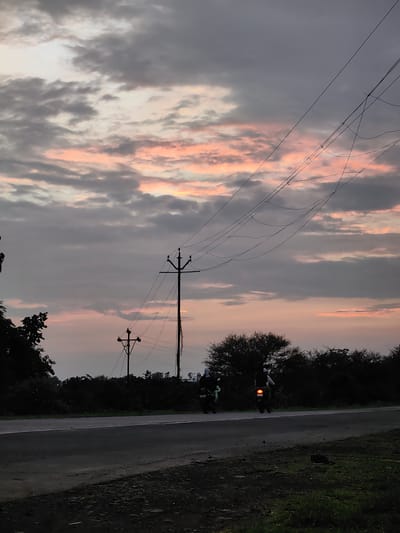 In Dattapur, India, at dusk, two individuals are seen riding motorcycles on a paved road. The sky is filled with dramatic clouds, tinged with shades of pink and grey, indicating the setting sun. Overhead, a complex network of power lines stretches across the frame, supported by utility poles. To the left, silhouetted trees line the roadside, and on the right, dense foliage creates a dark backdrop. The overall scene captures a moment of daily life against a striking natural sky.