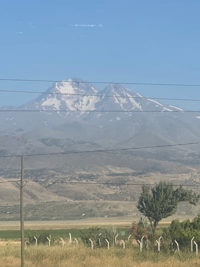 The image is a landscape photograph taken in the city of İncesu, Turkey, depicting a view of a snow-capped mountain in the distance. The mountain has a rugged texture and is partially obscured by haze. In the foreground, there is a dry, grassy field with a few sparse trees and a fence made of white posts. Power lines run horizontally across the frame, obscuring parts of the mountain and sky. The sky is a clear blue with some faint streaks of light that might be lens flare or atmospheric phenomena. The overall scene suggests a rural or semi-rural environment with a prominent natural landmark.