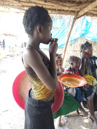 This image captures a group of people, primarily children, gathered under a makeshift shelter in Otulu, Nigeria. The shelter appears to be constructed from natural materials like wood, straw, and blue plastic sheeting. The scene suggests a moment of respite or a mealtime.
In the foreground, a young girl in a yellow and black patterned top and black skirt is shown from the side, eating from her hand. Behind her, two other children are visible. One child, looking directly at the camera with a wide-eyed expression, is also bringing food to their mouth. Another child, to the right, is looking away from the camera. A woman is seated further back, wearing a dark patterned top and touching her ear. She appears to be observing the scene.
A bright red and pink circular object, possibly a mat or a large dish, is positioned behind the girl in the foreground. In front of the seated woman and children, an orange and yellow plastic plate holds a small portion of what appears to be cooked food, possibly rice or pasta. A yellow plastic bag is also visible near the plate.
The lighting suggests it is daytime, with bright sunlight filtering through the shelter. The ground is dusty and uneven, consistent with an outdoor setting. The overall atmosphere is informal and appears to be a domestic or community gathering. No visible text is present in the image.