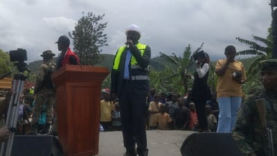 A man wearing a white hardhat and a yellow safety vest is speaking at a podium. He is standing in front of a crowd of people, many of whom are children. The crowd is standing on a platform made of concrete blocks. There is a large wooden podium in front of the man. To the left of the man, a person in a camouflage uniform is standing behind the podium. There is a camera on a tripod to the left of the podium. There is a truck behind the podium. There are banana trees and a hilly landscape in the background. The scene is likely taking place in Sake, Democratic Republic of the Congo, based on the provided coordinates.