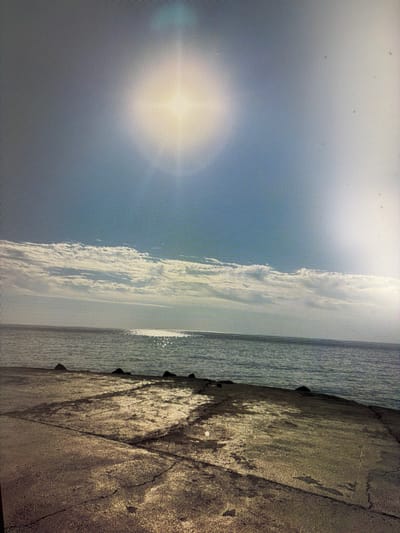 The image depicts a wide, static shot of a body of water meeting a concrete shoreline under a bright, partly cloudy sky. The sun is high, casting a bright glare on the water's surface and creating a hazy effect in the sky. The foreground is dominated by weathered, cracked concrete, suggesting an industrial or public access area. Dark, irregularly shaped rocks are visible along the edge of the water. The water itself appears calm, with subtle ripples reflecting the sky. The clouds are arranged horizontally, with a thin band of white clouds stretching across the horizon, contrasting with the clear blue sky above. There are no people or visible signs of activity in the scene. The location is specified as Mirnoye, Ukraine.