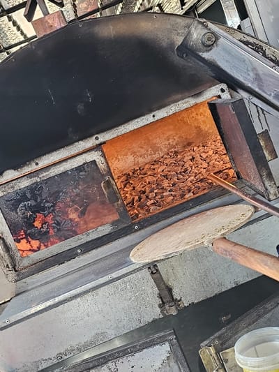 The image displays an angled, close-up view of a traditional baking oven in Boufakrane, Morocco. The oven's exterior is constructed from worn metallic panels, with a dark, curved section overhead. Inside, the left chamber contains active glowing embers and charred fuel. The right chamber features a substantial bed of hot, reddish-brown refractory material, radiating an orange glow. A round, light-colored flatbread dough is positioned on a long, wooden peel, extending into the hot right chamber. A copper-toned metal rod is also visible within this chamber, near the dough. A white plastic container is partially discernible in the lower right foreground.