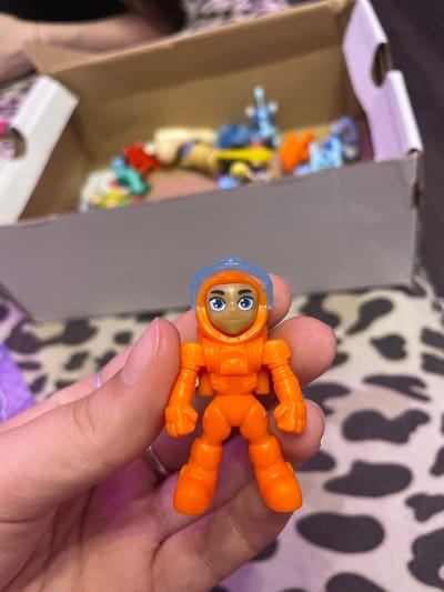 A close-up shot captures a small, orange astronaut figurine being held between a person's thumb and index finger. The figurine has a blue helmet with a clear visor, and its face is painted with blue eyes and a light brown skin tone. The astronaut's suit features details like a chest plate, arm joints, and articulated legs. In the blurred background, a cardboard box is visible, containing various other small toys and objects, some of which appear to be colorful figurines and building blocks. The patterned surface beneath the box and the fingers suggests a textile, possibly a blanket or clothing, with a cow-print or similar abstract design. The lighting is dim, casting soft shadows and highlighting the textures of the figurine and the skin. The scene is devoid of any recognizable landmarks or elements that would indicate a specific location within Ladozhskii Trudposelok, Russia.