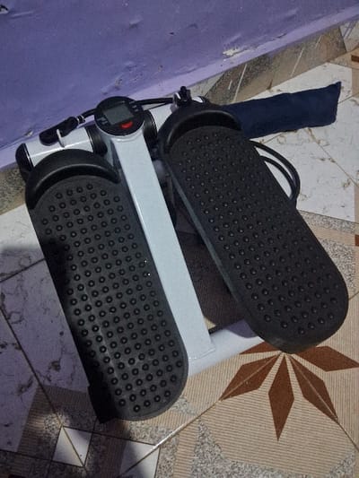 The image displays an exercise mini-stepper placed indoors on a tiled floor, next to a wall. The stepper features a white metal frame supporting two prominent black foot pedals. Each pedal is textured with an array of small, raised circular nubs designed to provide grip. A small digital display unit, which includes a red button or indicator, is visible on the central white frame, accompanied by some thin wires or cables. Behind the upper section of the stepper, a dark blue fabric object, possibly a mat or a protective cover, is partially seen.
The floor beneath the stepper is covered with ceramic tiles that exhibit varied patterns. Some tiles are light beige and gray with a marble-like texture, while others feature a distinct brown and white starburst geometric design. To the left of the stepper, a light purple-colored wall is visible, showing signs of wear with patches where the paint appears to be peeling or chipped.
No people are present in the image, and the exercise equipment is stationary, indicating it is either stored or not currently in use. The indoor setting offers no clues regarding the time of day or outdoor weather. Any text or numerical readings on the digital display are too indistinct to be legible. The scene is captured from a slightly elevated and angled perspective.