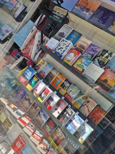 The image captures a brightly lit display window of a bookstore, likely in Antony, France, given the context. The window showcases a dense arrangement of books and some fabric items. The books are mostly paperbacks with colorful covers, arranged in tiered shelves. Some book titles are partially visible, including "Michael Connelly," "Le Voyage de Mille Lieux," and "Les Crimes de la Maison Bleue." Interspersed among the books are what appear to be decorative fabric items, possibly scarves or bags, with floral and illustrated patterns.

The lighting is intense, suggesting artificial illumination within the store, and the glass reflects some of the exterior environment, including trees and sky. There are no people visibly present in the immediate foreground or interacting with the display. The overall impression is of a well-stocked, vibrant retail space.