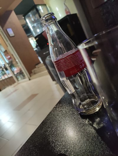 The image depicts an empty clear glass Coca-Cola bottle with a red label, placed on a textured dark surface, possibly a table. The bottle is angled and tilted towards the right of the frame. A portion of a reflective surface, possibly another glass, is visible to the right of the bottle. The background is blurred, suggesting an indoor environment with some lighting. The flooring in the background appears to be tiled. Elements of a display case with illuminated items are visible in the upper left portion of the background, along with a muted brown wall. The scene is consistent with a casual dining or bar setting, potentially within Madan, Bulgaria, though no specific landmarks are identifiable.