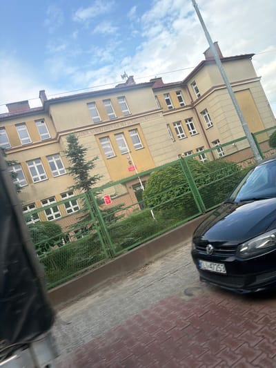 The image shows the exterior of a school building with a sign that reads "SZKOŁ SAMORZĄDOWYCH NR 2 IM. TADEUSZA KOŚCIUSZKI". A black car is parked in front of a green metal fence, partially obscuring the view of the building. The building appears to be old, with yellow-colored walls and red-tiled roofs. There are trees and bushes in front of the building. The sky is cloudy. The ground is paved with bricks. The image is taken from a slightly tilted angle, suggesting it might have been taken from a moving vehicle. The location is the Tadeusz Kościuszko Primary and Middle School No. 2 in Limanowa, Poland.