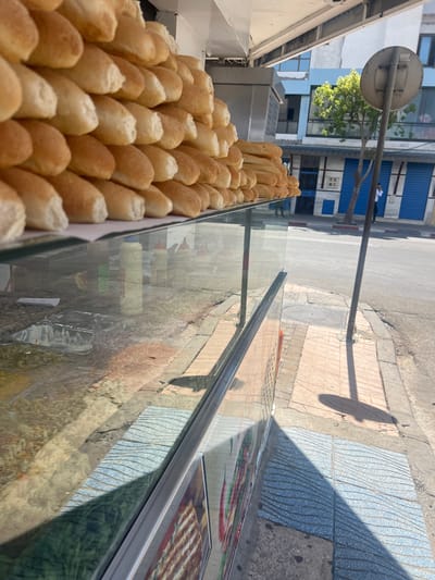 The image captures a street scene in Martil, Morocco, focusing on a display of bread rolls. The rolls, uniform in size and shape, are densely packed in a tray or container. The display appears to be part of a mobile or street vendor setup, with a glass or transparent barrier separating the rolls from the outside environment. This barrier provides a partial reflection of the surroundings, including street features, a building facade, and presumably other items behind the photographer. The backdrop of the scene includes a street with various textures and patterns in the pavement, a building with blue shutters or paneling, and a street sign. A pole extends into the scene, casting a shadow on the pavement. The overall impression is of a typical street food scene in Martil.