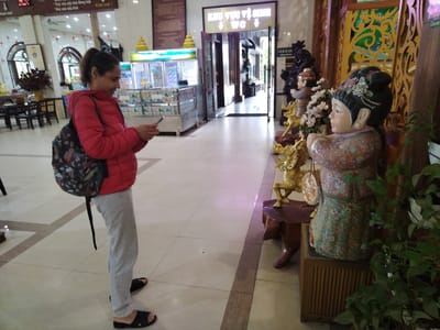 A woman stands in a brightly lit indoor space, possibly within a temple or religious building. She is looking at her phone, dressed in a red jacket, gray sweatpants, and dark sandals. She also wears a backpack.
To her right are decorative statues, including a statue of a girl and another statue of a figure riding a golden horse. To her left is a refrigerated display case with items inside.
Behind the woman, in the center of the image, is a doorway marked "KHU VỰC VỆ SINH WC" in Vietnamese, indicating the location of restrooms. The floor is tiled and the space seems well-maintained. The location is in Chu Văn An Ward, Vietnam.
