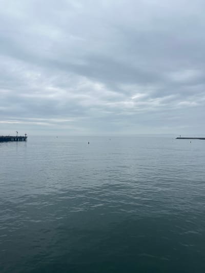 The image displays a calm ocean vista under an overcast sky in Santa Barbara, California, United States. The viewpoint is from the Santa Barbara coastline, looking seaward into the Santa Barbara Channel. To the left, a portion of Stearns Wharf is visible, characterized by its wooden pilings, deck, and several light poles, one bearing a red marker. To the right, a stone breakwater extends into the water, culminating in a navigational aid or light structure. The mid-ground ocean surface features several small, red and orange cylindrical buoys. The sky is uniformly gray with layered clouds, and the water is dark gray-blue with subtle ripples. No individuals or active vessels are present.