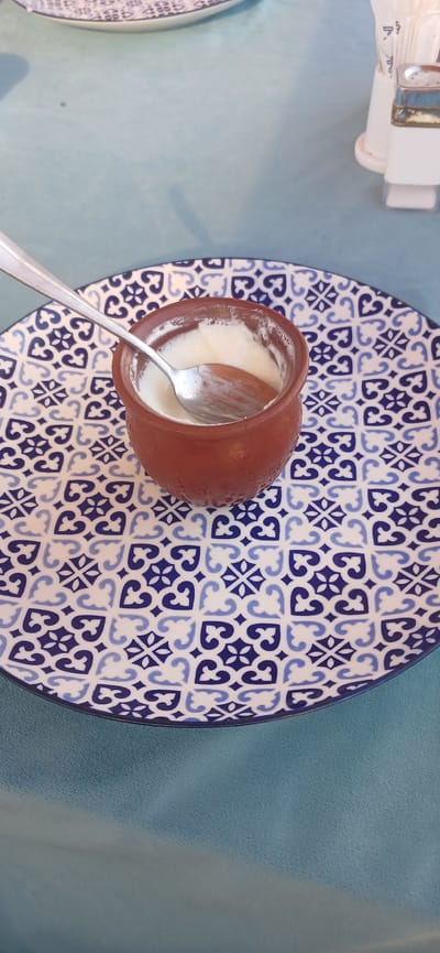 This media file shows a close-up, overhead view of a brown clay pot filled with yogurt, placed on a decorative blue and white patterned plate. A silver spoon is resting inside the pot, slightly submerged in the yogurt. The plate itself is positioned on a light blue, textured surface, possibly a tablecloth or a placemat. To the upper left of the frame, a white object with some metallic accents is visible, which appears to be a napkin holder.
The scene suggests a meal is being consumed or has recently been eaten, given the presence of the yogurt and spoon. The patterned plate and clay pot hint at a rustic or traditional culinary setting. The lighting appears to be natural daylight, casting soft shadows, suggesting it could be daytime. There are no visible people, and the focus is entirely on the food item and its presentation. No text is discernible in the image. The context of Sumqayit, Azerbaijan, suggests this might be a local dish or a meal served in a restaurant in that region.