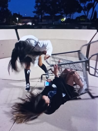An outdoor scene at dusk or night in the city of Kukcha, Uzbekistan, depicts two individuals on a light-colored paved surface. One individual, wearing a white long-sleeved top, dark shorts, and dark knee-high socks with white bows, is bent forward with her head obscured by long dark hair, which appears in motion. The second individual is lying supine on the ground, partially covered by an overturned metal shopping cart. This individual wears a dark long-sleeved top, dark shorts, and white sneakers, with long dark hair spread on the pavement. Her hands are raised and open. A light-blue mobile phone rests near her left hip. In the background, a low concrete barrier is visible, with trees and artificial lights against a dark blue sky.