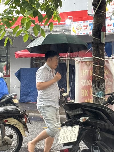 The scene depicts a street in Ngũ Hành Sơn Ward, Vietnam, under rainy conditions. An adult male, of apparent East Asian ethnicity, walks barefoot from left to right, shielding himself with an open dark-colored umbrella. He is dressed in a light-colored, short-sleeved collared shirt with a subtle pattern and light-blue denim shorts. His right hand is partially obscured, raised near his chest.
The foreground includes the rear portion of a light-colored motorcycle on the left and the rear of a black motorcycle on the right, bearing Vietnamese license plate '37-B2 790.63'. A tree trunk, partially wrapped in white material, stands vertically on the right. Green foliage from a tree overhangs the upper left.
The background shows commercial establishments with a prominent red awning, a blue tarp covering an unseen object, and various signs with Vietnamese text, including 'ĐỒNG GIÁ' (Same Price) and 'TRÀ SỮA' (Milk Tea) visible on one sign, and fragments of text 'IHT U' on another. The ground surface is visibly wet from rain.