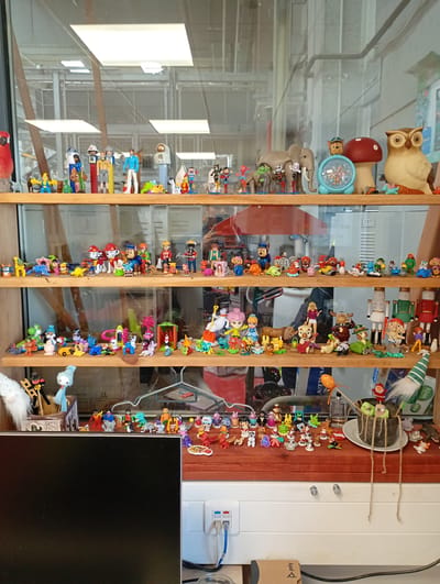 This image captures a multi-tiered wooden and glass display case filled with a vast and eclectic collection of small figurines and toys. The setting appears to be an indoor environment, possibly an office or workshop, with large windows and industrial-style ceiling structures visible through the glass shelves as reflections and background elements.
The shelves are densely packed with a diverse array of figures. The top shelf showcases characters such as R2-D2, stormtroopers, Chewbacca, and Luke Skywalker from Star Wars, various Playmobil figures including a firefighter and an astronaut, superheroes like Spider-Man, and several Pez dispensers. Prominent items on this shelf include a large grey elephant, a light blue alarm clock topped with Paw Patrol's Chase, a decorative red mushroom, and a substantial wooden owl figure. A red parrot sits on the far left.
The middle shelves feature a continuation of this varied collection, with numerous Paw Patrol figures, Smurfs, Super Mario's Yoshi, more Playmobil characters (such as a cowboy, doctor, and knight), an assortment of animal figures (giraffes, dinosaurs, dogs, cats, bears), and small vehicles. Also present are decorative nutcracker figures, various dolls resembling Disney princesses and a Barbie-like figure, and a range of gnomes and fantastical creatures.
The bottom shelf, partially covered by a black computer monitor on the left, displays additional figurines including superheroes, animals like penguins and gorillas, and more Playmobil characters. To the right, a small potted plant with string adornments sits beside a white ceramic plate. A fluffy white gnome and a light blue doll are also visible.
In the foreground, a black computer monitor occupies the lower-left section. To its right, a white wall features power outlets and network cables (blue and grey) plugged in. A small brown cardboard box with the word "poly" printed on it rests near the cables. A white sticker with the number "237" is visible on the glass in the background. The collection is a static display within an indoor setting, in Kabile, Bulgaria.