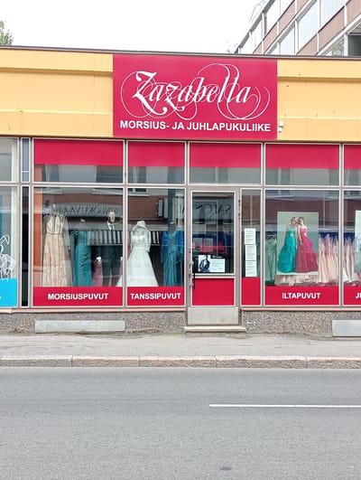 The image shows the storefront of "Zazabella", a formal wear shop in Kuopio, Finland. The shop's facade is painted yellow and red, with the store name prominently displayed in white script on a red banner. Below the banner, the text "MORSIUS- JA JUHLAPUKULIIKE" indicates the types of garments sold: wedding and party dresses. The storefront features large display windows showcasing various dresses on mannequins and racks. Some of the displayed items include a beige, sequined gown, a teal-colored dress, a groom's suit, a white wedding dress, and a blue formal dress. The window on the right displays a poster of two women in long, colorful evening gowns, with more evening gowns visible on racks behind them. Text labels like "MORSIUSPUVUT" (wedding dresses), "TANSSIPUVUT" (dance dresses), and "ILTAPUVUT" (evening gowns) are visible on red strips below the window displays. A glass door with a "SALON" sign is visible to the left of the evening gown display. The shop is situated on a street with a sidewalk and a road in front of it. The building appears to be a commercial establishment with multiple floors, as indicated by the windows visible above the storefront. The overall scene depicts a well-lit day in an urban environment.