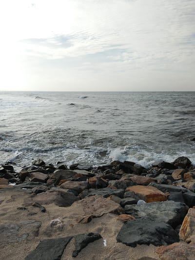 The image depicts a rocky shoreline with the ocean in the background. The sky is overcast with scattered clouds, and the sea is choppy with small waves breaking near the shore. Large, dark rocks are scattered along the edge of the water, and the foreground consists of sand and smaller rocks. There are no people or discernible man-made structures in the image, suggesting a natural, coastal environment. The scene is likely a beach or a rocky coast within the city of Phú Thủy, Vietnam.