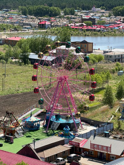 The image, taken from a high vantage point, presents an outdoor amusement park and a residential area under bright daylight. A prominent pink Ferris wheel, equipped with multi-colored gondolas (red, green, and yellow), dominates the center. Below it, within the amusement park, a colorful carousel and other rides are visible on artificial green turf. Surrounding the park are various buildings, some with traditional Chinese architectural elements and others with more modern facades.

A calm body of water, possibly a river or canal, separates the amusement park from a village or town. This settlement features numerous houses, many distinguished by red or blue roofs, spread across the landscape and backed by dense green trees, suggesting a forested hill or mountain. Few people are discernible, with only faint figures on the carousel and near the rides, indicating a quiet period at the park.

Clear shadows confirm it is a sunny day, likely around midday. On a red banner hanging from a building on the right, Chinese characters are visible, partially obscured. Identifiable text includes "渔家" (yú jiā - fisherman's home/restaurant) and phrases such as "特色江鲫鱼" (tèsè jiāng jìyú - specialty crucian carp) and "酒店" (jiǔdiàn - hotel/restaurant), suggesting a restaurant or hotel specializing in local fish. A black SUV and other vehicles are parked nearby. The scene depicts a leisure area within Jiejinkou, China.