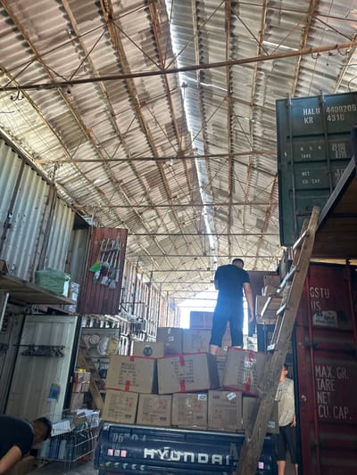 The image depicts an indoor storage area, possibly a warehouse or a loading bay, in Kara-Suu, Kyrgyzstan. The space is characterized by a corrugated metal roof supported by a metal framework, with visible light filtering through gaps. Large shipping containers, predominantly green and red, are stacked and form part of the structure. In the foreground, a man in a dark t-shirt and black shorts stands on top of a stack of cardboard boxes on the back of a truck. He appears to be looking down. Another person, wearing a light-colored t-shirt and dark shorts, is partially visible on the right, standing near the boxes and a wooden ladder leaning against a container. Various cardboard boxes, some with shipping labels and markings, are stacked on the truck and in the surrounding area. Cleaning supplies, including mops and brooms, are hung on a red container door. Bags and other items are placed on shelves. The overall impression is one of a busy, functional storage and loading environment.