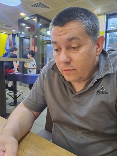 A man with gray hair and an Adidas gray polo shirt is sitting at a wooden table. The man is inside a restaurant, with customers visible in the background. The restaurant has blue chairs and yellow booths. The location is Bel'kovo, Russia.